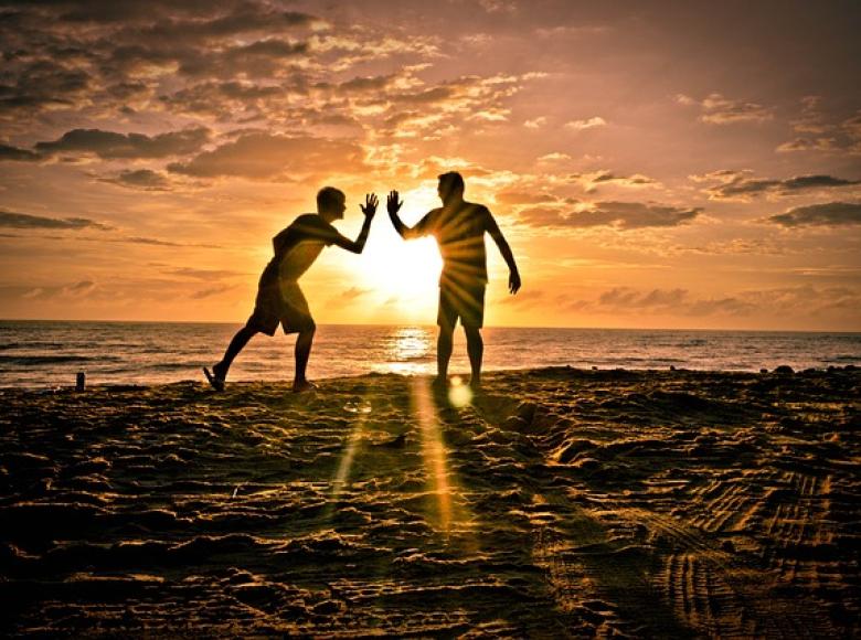 Two friends giving high fives to each other on a beach with sun setting in the background