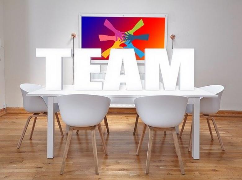 The word TEAM in large white letters on a white table with four white chairs, over a hardwood floor, with white walls in the background and a colorful picture on the wall of hands coming together 